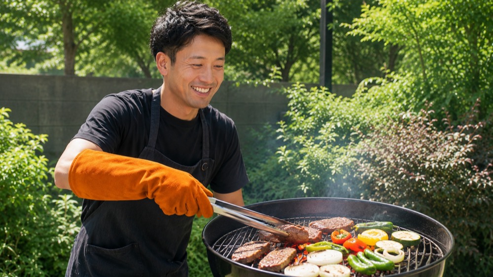 オレンジの耐熱グローブをはめた手で、炭が整ったグリルで肉を焼いている満足そうな男性