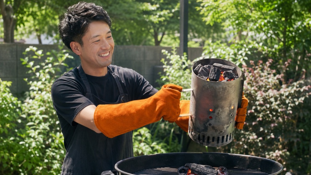 庭のBBQで熱い火起こし器をオレンジの耐熱グローブで持ち上げ炭を移す男性