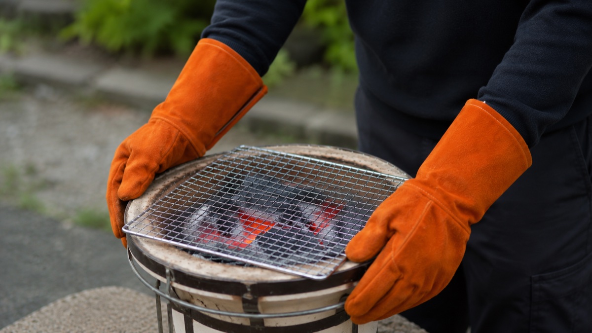 屋外の七輪そばでオレンジの耐熱グローブをはめた手が炭の位置を調整している